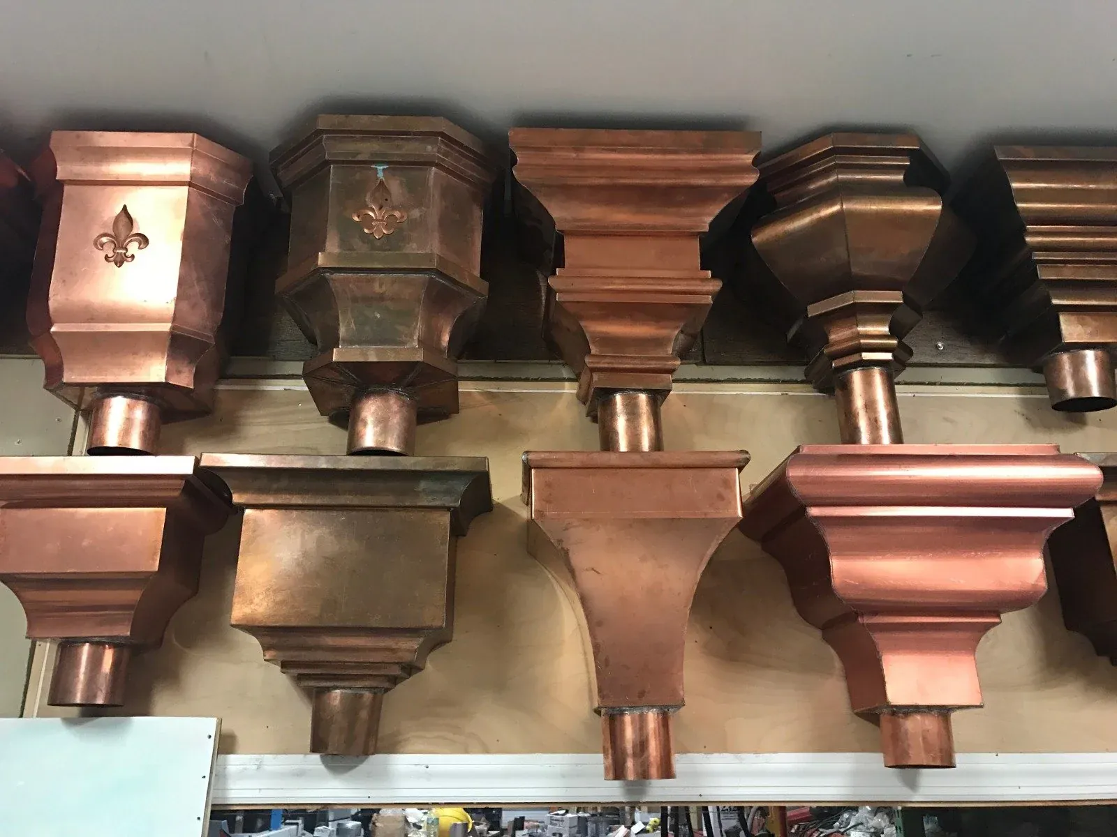 Rows of custom copper leader heads in different shapes, some with a fleur-de-lis, displayed on a shop wall