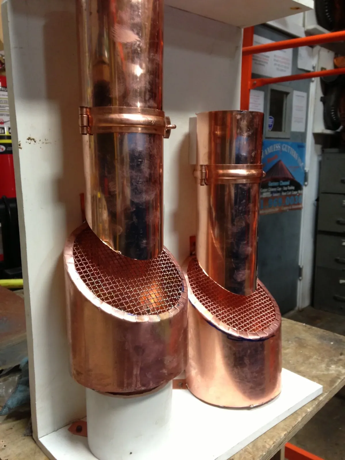 Two copper leader pipe elbows with mesh strainers mounted on a display board