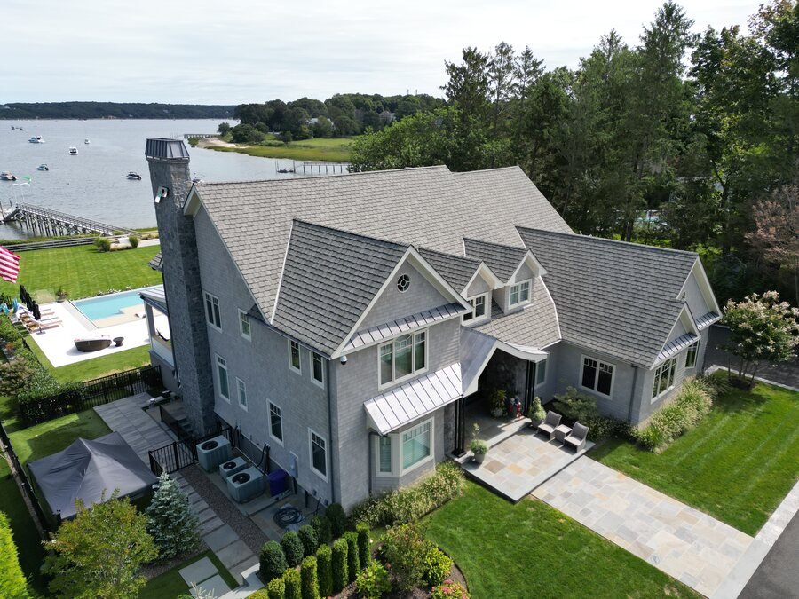 Aerial view of a gray shingled waterfront house with a stone chimney, standing-seam metal awnings and a pool