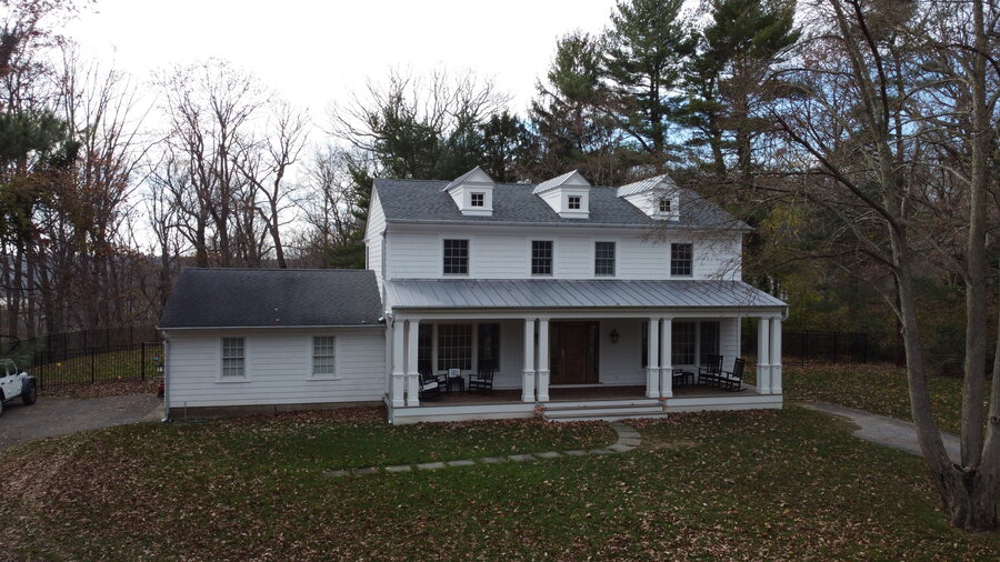 White two-story farmhouse-style home with a gray shingle roof, a metal porch roof and dormers, among fall trees
