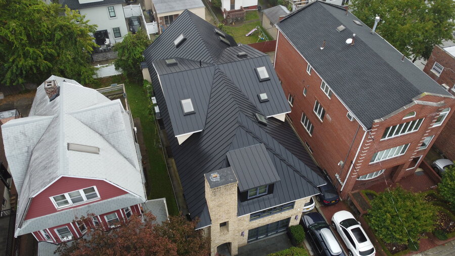 Top-down view of a dark metal standing-seam roof with skylights between a brick building and a red house