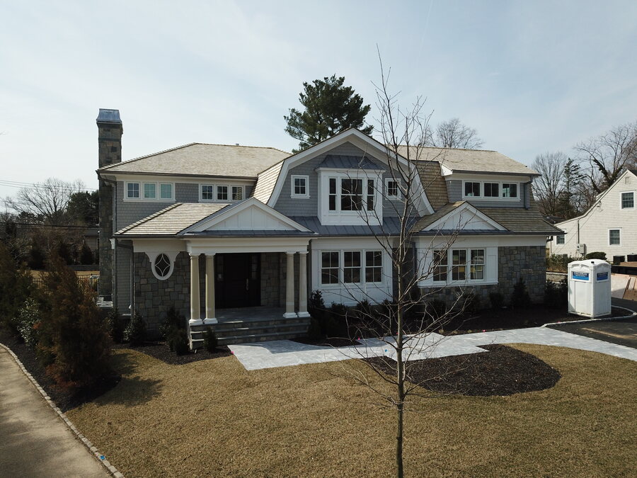 Front view of a gray shingle-style house with stone chimney, columned entry, oval window and a young tree in the lawn