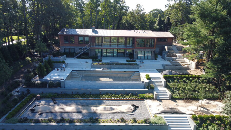 Modern hillside house with a metal roof and glass walls above terraced stone steps, a pool and landscaped gardens