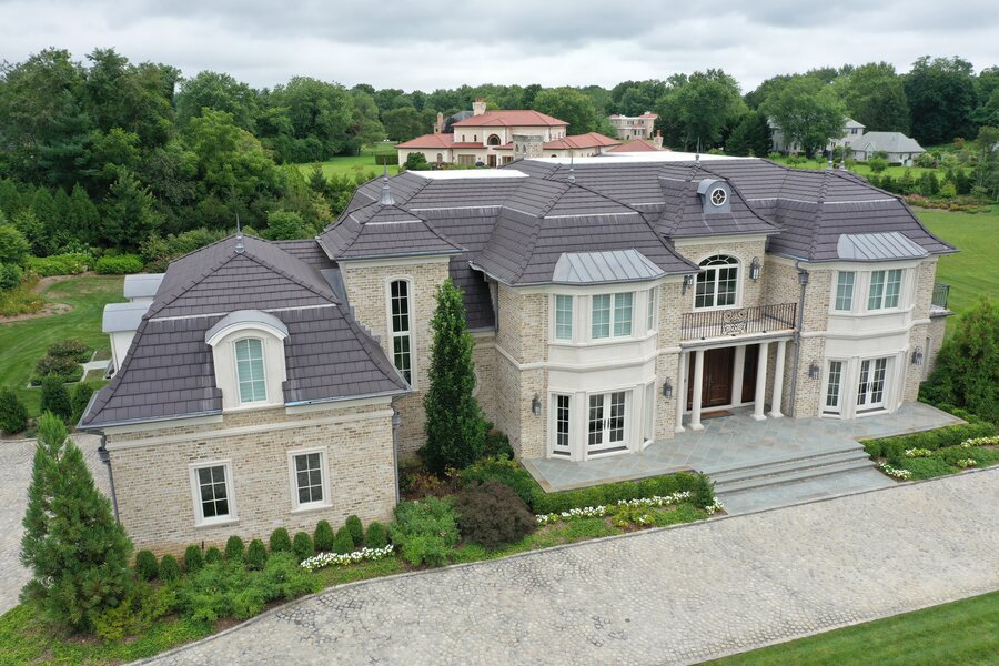 Aerial view of a large limestone-colored mansion with a dark tile roof, metal-roofed bays and a stone terrace