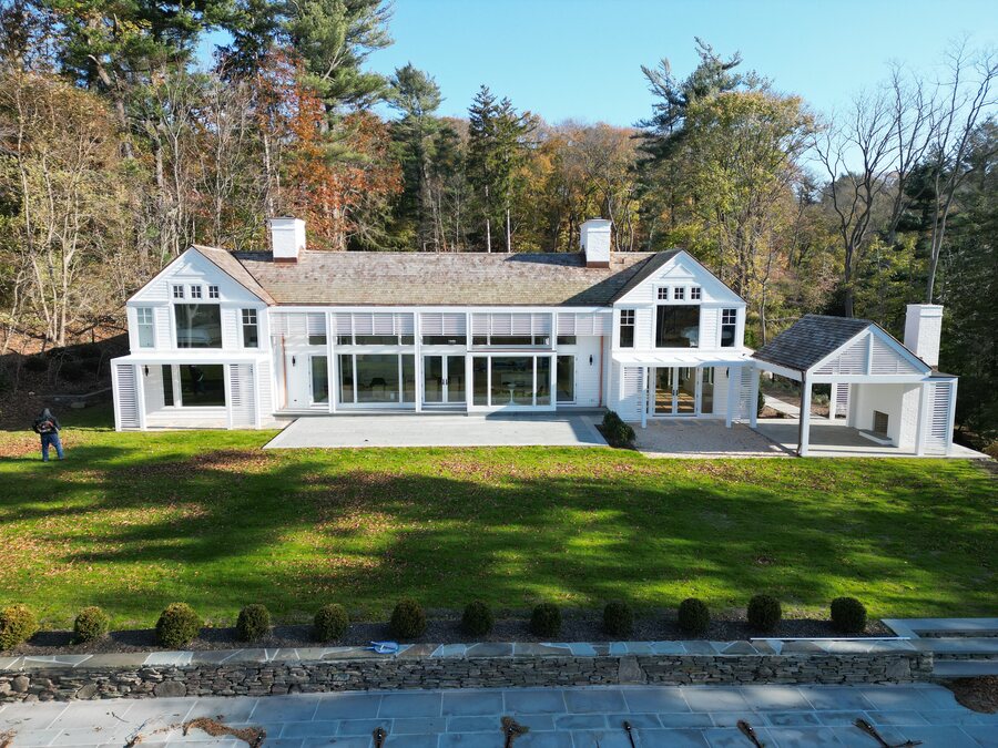 White house with tall glass doors, louvered shutters, two chimneys and a covered pavilion beside a stone patio
