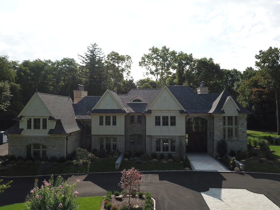 Stone and cream-siding house with gabled dormers, a dark shingle roof and an arched entry beside a paved driveway