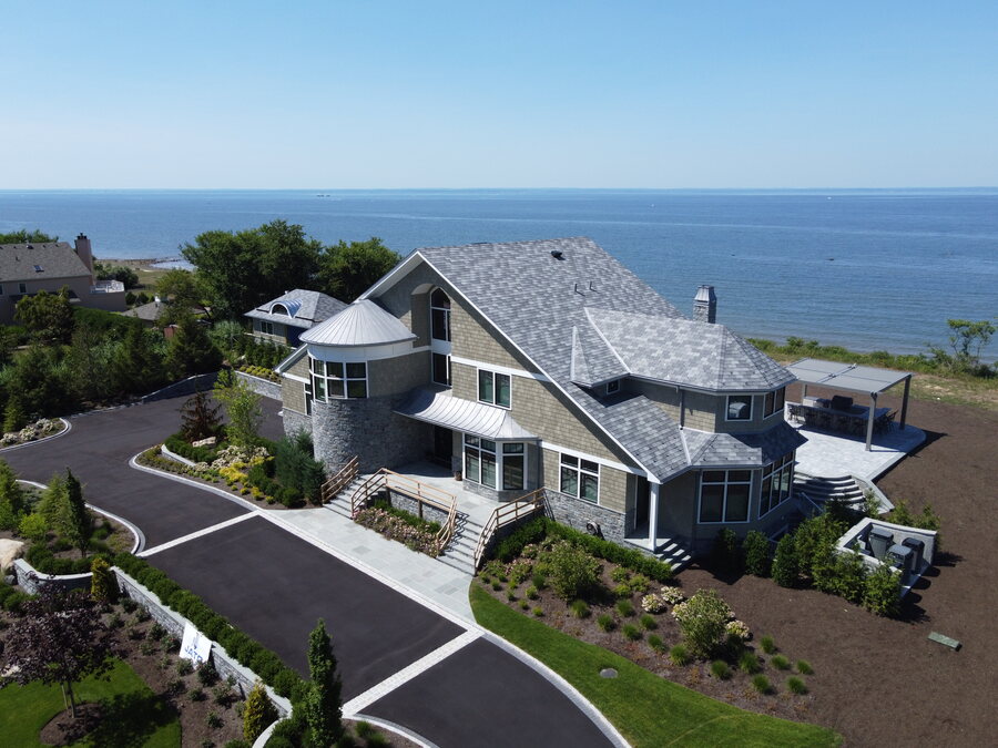 Aerial view of a shingled house on a bluff over open water, with a round metal-roofed turret and a curving driveway