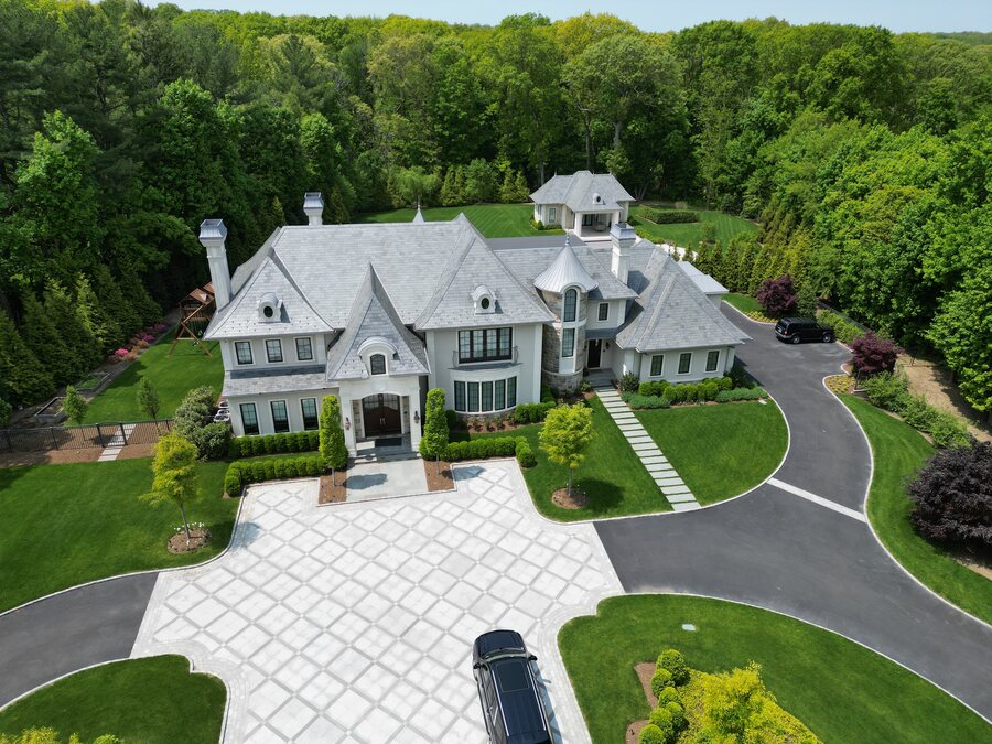 Aerial view of a gray stone and slate-roofed mansion with a round turret, a paver motor court and wooded grounds