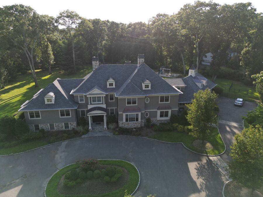 Aerial view of a gray shingle-style house with dormers and a slate-look roof beside a circular driveway