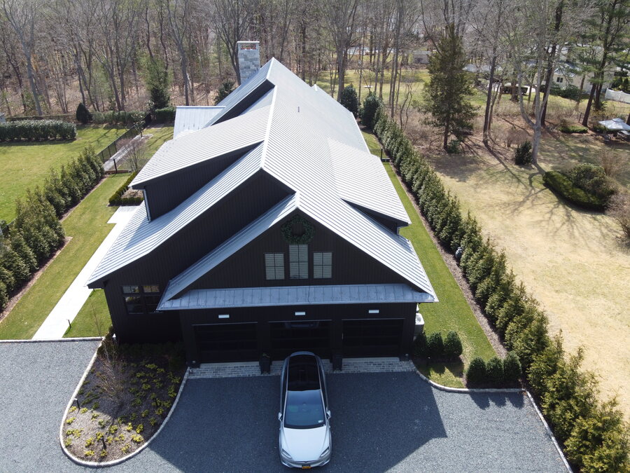 Top-down view of a dark house with a light standing-seam metal roof and a gravel motor court