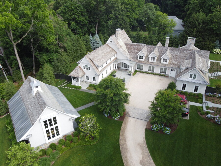 Aerial view of a white shingle-style estate with a cedar roof and a gray metal-roofed outbuilding