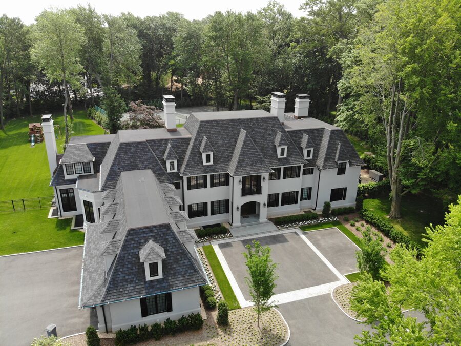 White house with a dark slate-look roof, multiple dormers and a paved motor court