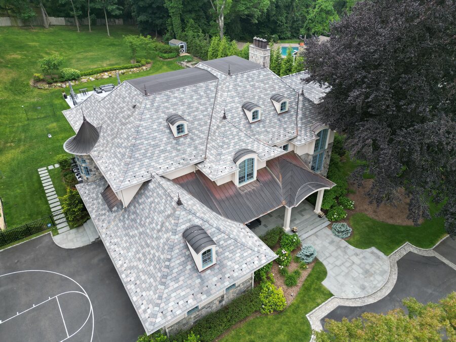 Aerial view of a gray slate-look roof with arched dormers and a curved copper porch roof