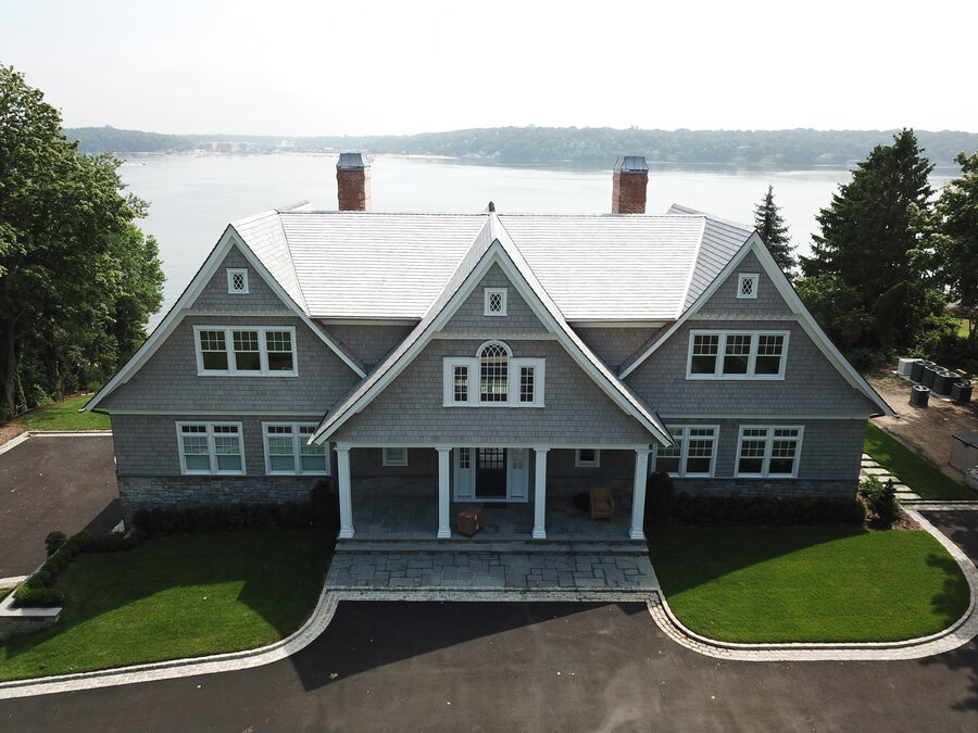 Waterfront gray-shingle house with a light slate-look roof, two brick chimneys and a covered entry